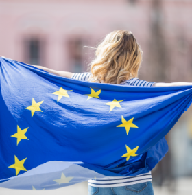 A young woman with her back to the camera, holding a fluttering European flag behind her back
